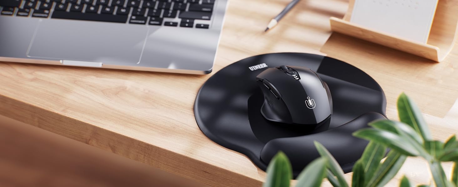 Black circular computer mouse on wooden desk, next to partial view of laptop keyboard and plant leaves.