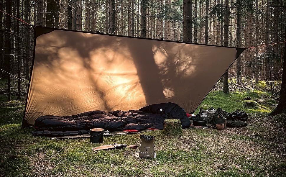 Tan tarp shelter set up in a forest. Sleeping bag and camping gear visible underneath. Sunlight filtering through trees.