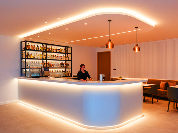 Modern bar interior with curved illuminated counter, backlit bottle shelving, and copper pendant lights. Features warm ambient lighting and lounge seating.