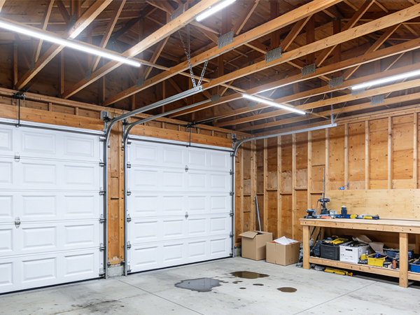 Empty garage interior with white overhead door, wooden wall framing, and workbench with tools, illuminated by warm overhead lighting.