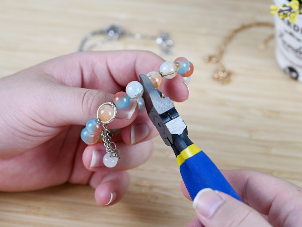 Hands manipulating jewelry with pliers. Colorful beads and charms visible, including blue and peach tones. Wooden surface in background.