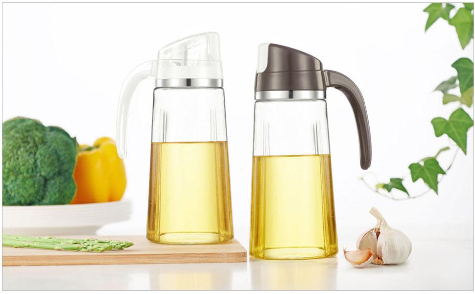 Two glass oil dispensers with automatic flip-top lids, one clear and one brown. Containers filled with yellow cooking oil, displayed on wooden surface with vegetables.