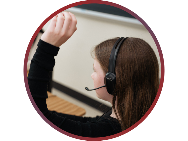Image of a young girl wearing a headset sitting at a desk and raising her hand.