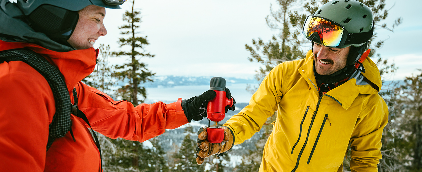 Two skiers in bright jackets exchanging a beverage on snowy slope. One wears orange, the other yellow, both with ski goggles.