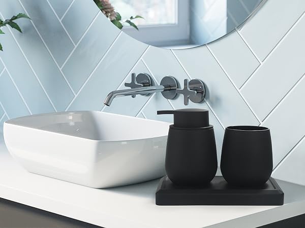 Modern bathroom vanity with white vessel sink, wall-mounted faucet, and black ceramic accessories on a tray. Herringbone tile backsplash and circular mirror visible.