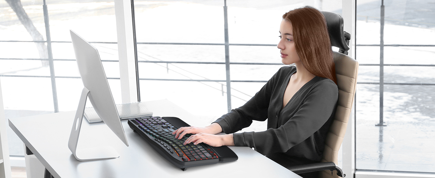 Woman in business attire sitting at desk working on desktop computer with monitor in bright office space with floor-to-ceiling windows overlooking cityscape.