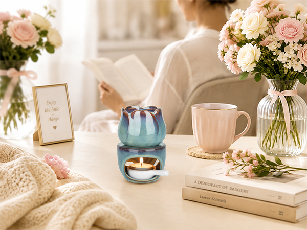 Teal glass decorative diffuser or candle holder with geometric pattern, positioned on white surface among pink mug, books, and fresh flowers in clear vases. Soft neutral home décor styling with natural lighting.