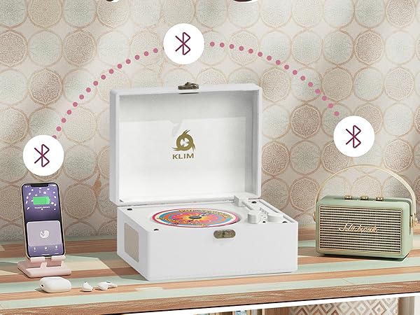 White vintage-style record player with Bluetooth connectivity icons shown, displayed with speakers and phone on patterned fabric surface.