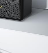 Close-up of a black electronic device with a perforated speaker grille, partially visible against a light-colored surface.