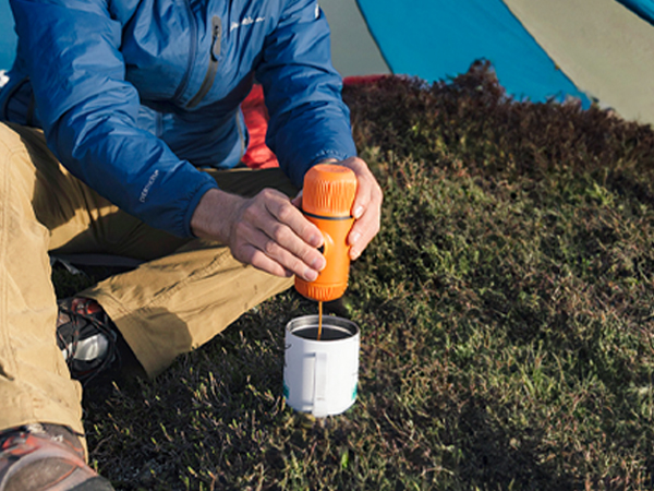 Person in blue jacket and tan pants using handheld orange power tool to mix contents in white container outdoors.