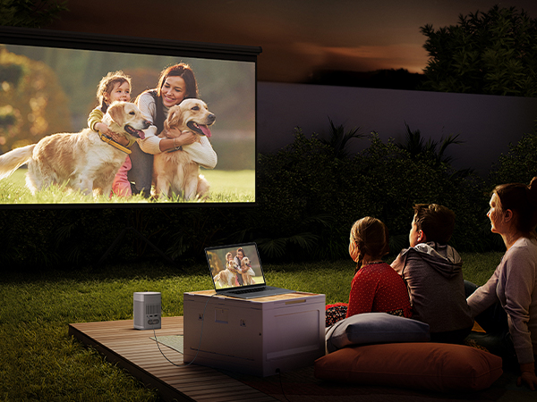 Outdoor movie screening setup showing projected image on white screen, with viewers sitting on cushions in darkened backyard setting.