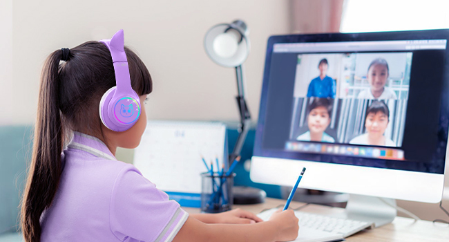 Purple wireless headphones worn by person attending video call on desktop computer. User writes notes while participating in multi-person virtual meeting.
