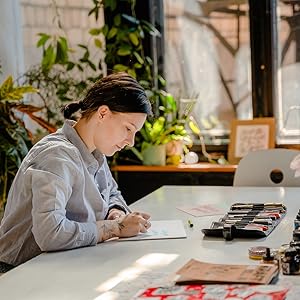 Person sitting at a desk with art supplies, including paintbrushes and paints. Surrounded by plants in a well-lit workspace with a laptop visible.