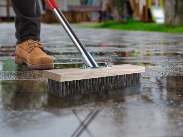 Large push broom with stiff bristles being used to sweep water on wet concrete surface, shown from side angle with partial view of work boot.