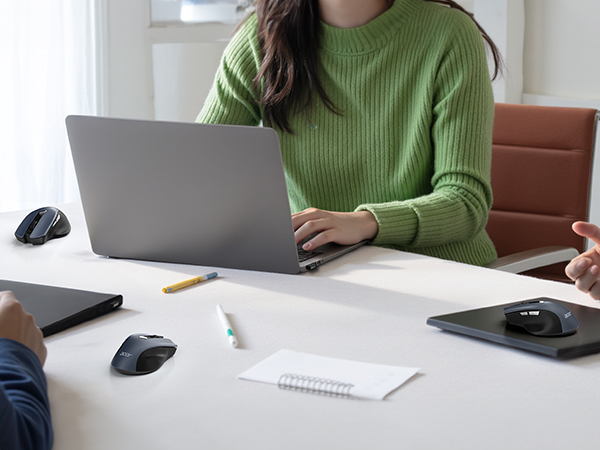Person in green sweater using laptop at white desk. Office supplies and wireless mice visible. Brown chair in background.