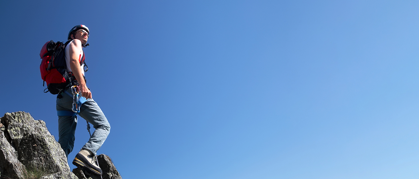 Person wearing red backpack standing at edge of rocky mountain peak or cliff, viewed from side angle against cloudy sky background.