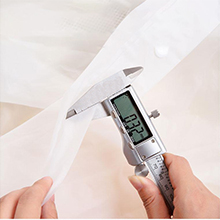 Hand holding digital caliper measuring thickness of white fabric or material.
