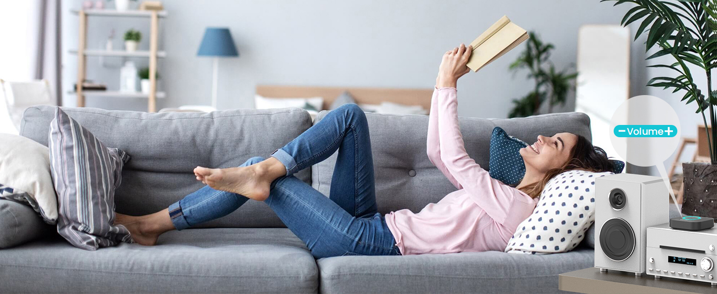 Lifestyle image showing someone relaxing on a grey couch with pillows, using a mobile device while lying down in a modern living room setting with shelving and plants.