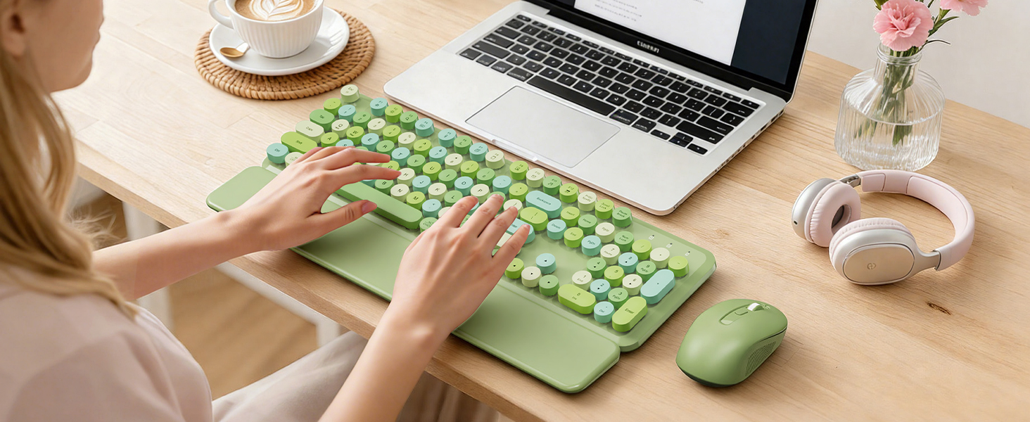 Person typing on green keyboard with wrist rest and mouse beside a laptop on desk