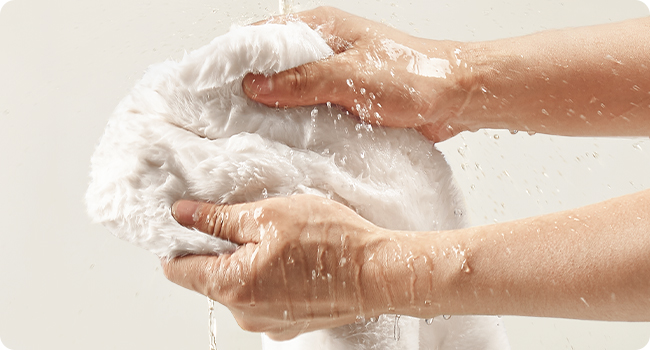 Hands lathering and rinsing a white cloth or sponge with water, demonstrating washing or cleaning action.