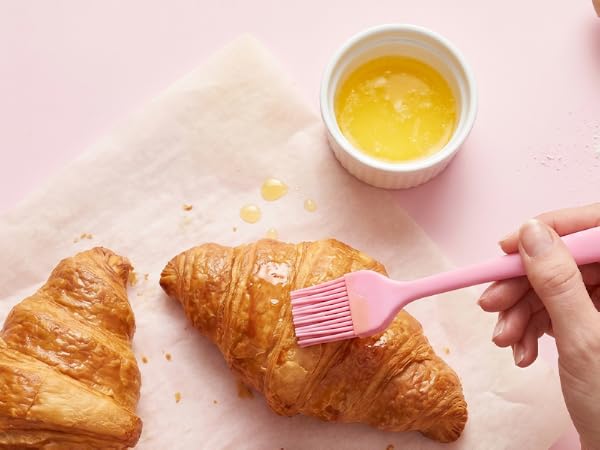 Pink silicone pastry brush spreading melted butter on croissants on pastel pink background