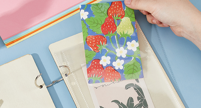 Colorful planner or notebook with metal rings, featuring a decorative cover insert with strawberry and flower pattern. Hands visible arranging pages.