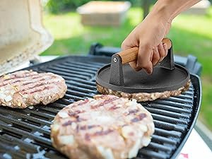 Hand using a cast iron grill press on burger patties cooking on a barbecue grill grate.