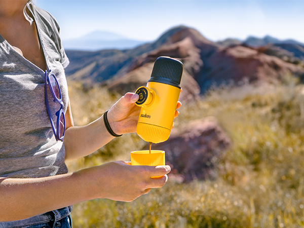 Yellow portable espresso maker being used outdoors against mountainous desert landscape. Person pouring coffee into small cup while hiking.