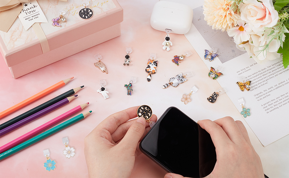 Crafting scene with colorful pencils, small butterfly and flower charms, and hands working with a black tool on a pink surface.