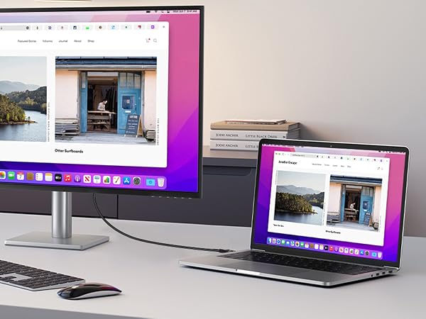 Desktop setup with large monitor and laptop displaying identical image of lakeside cabin. Wireless keyboard and mouse on desk. Books stacked in background.