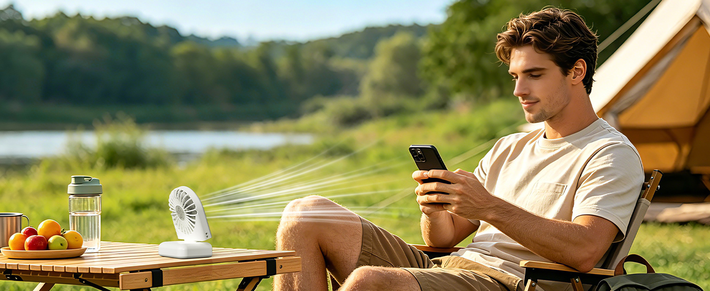 Man in tan shirt seated outdoors near tent and camping table, holding smartphone with lake and vegetation in background.