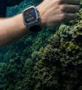 Close-up of smartwatch on wrist against lush green foliage background, showing dark watch face and band.