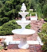 3 tier white outdoor water fountain with pineapple topper in a lush landscaped backyard garden