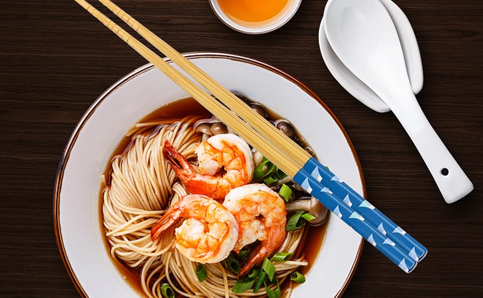 Bowl of noodle soup with shrimp, chopsticks, spoon, and small dish of sauce on wooden table. Noodles in broth with green onions and large cooked shrimp visible.
