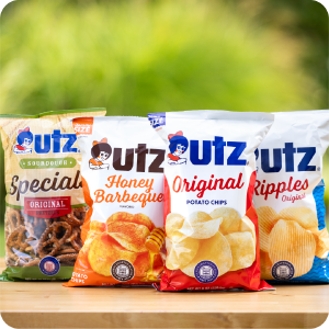 Four Utz snack bags on wooden surface with green outdoor background, featuring pretzels and chips.