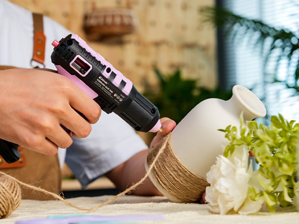 Black hot glue gun being used to apply adhesive to a white vase or container wrapped with twine, with greenery and flowers visible.