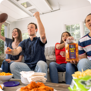 Four friends cheer on a couch, holding a football, Utz Party Mix, and snacks in a bright room.