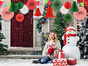 Christmas scene with decorated doorway, colorful paper fans, snowman, and wrapped gifts. Person sitting on snowy ground holding a red mug.