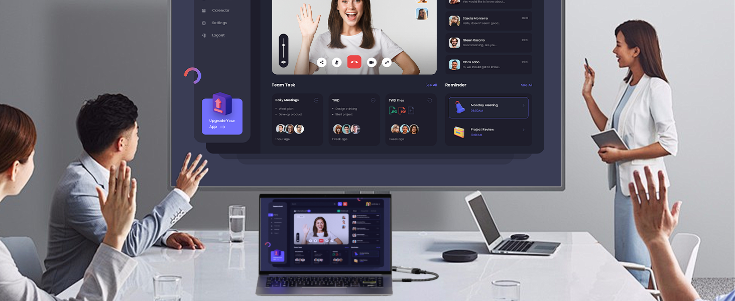 Video conferencing setup with large screen showing multiple participants. People in a room gesturing towards the screen. Laptop on table displaying related content.