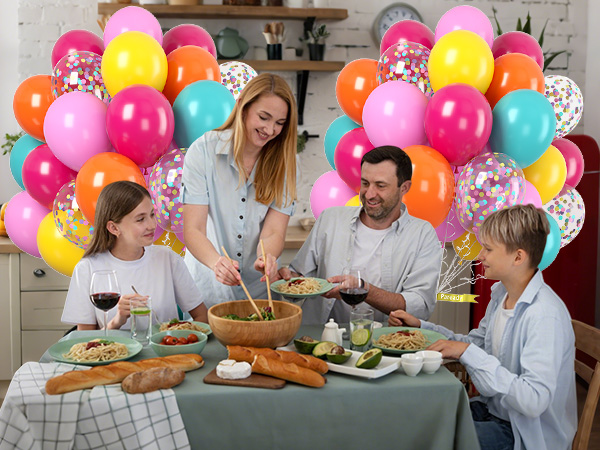 Dining table set with food and colorful balloons in background, featuring orange, pink, yellow, and blue balloons with confetti patterns.