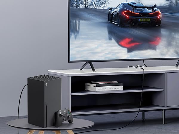 Modern gaming setup with large TV displaying race car, Xbox Series X console, controller on round table, and sleek entertainment center with shelves.