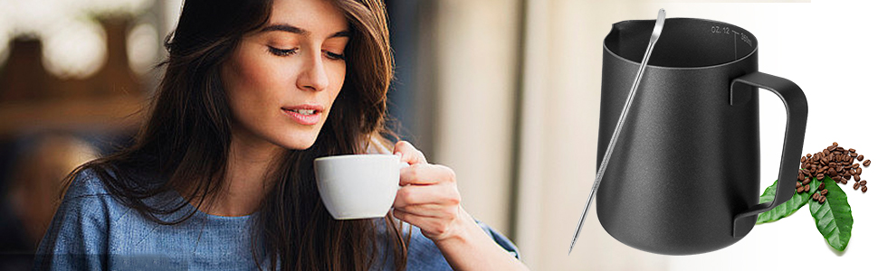Black stainless steel milk frothing pitcher with sleek design, positioned next to coffee beans and leaves. Woman drinking from white cup in background.
