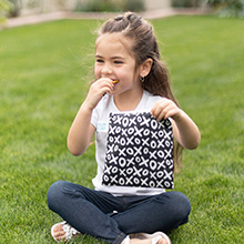 Girl sitting on a lawn and holding a black cloth snack bag while eating a pretzel
