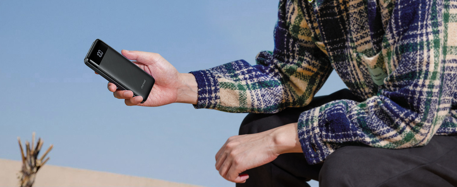 Person in plaid shirt sitting outdoors, holding a black smartphone in one hand against a natural background.