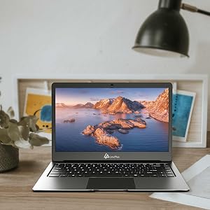 Laptop displaying coastal mountain scene on screen, positioned on desk with desk lamp and framed artwork visible in background.