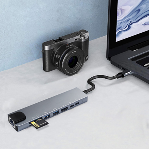 Compact digital camera connected to a laptop via a multi-port USB hub adapter on a white desk surface.