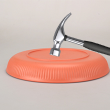Silver hammer striking an orange circular object with ridged edges. The hammer's head is in contact with the object's surface, suggesting impact or testing of material properties.