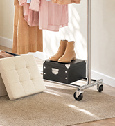 Black wheeled storage organizer with compartments on wooden floor next to beige cushions and home décor items.