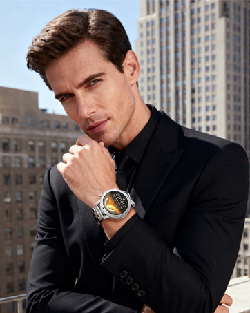 Close-up product shot showing luxury watch with metallic band against urban background. Subject wearing black suit jacket.