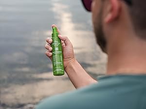 A man is holding a green bottle of Absolutely Natural Insect Repellent near a calm body of water.